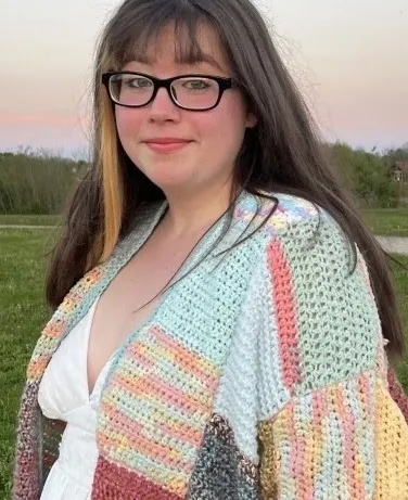 Grace Shields, smiling, with long dark hair and glasses, in a pastel crocheted cardigan outdoors at dusk.